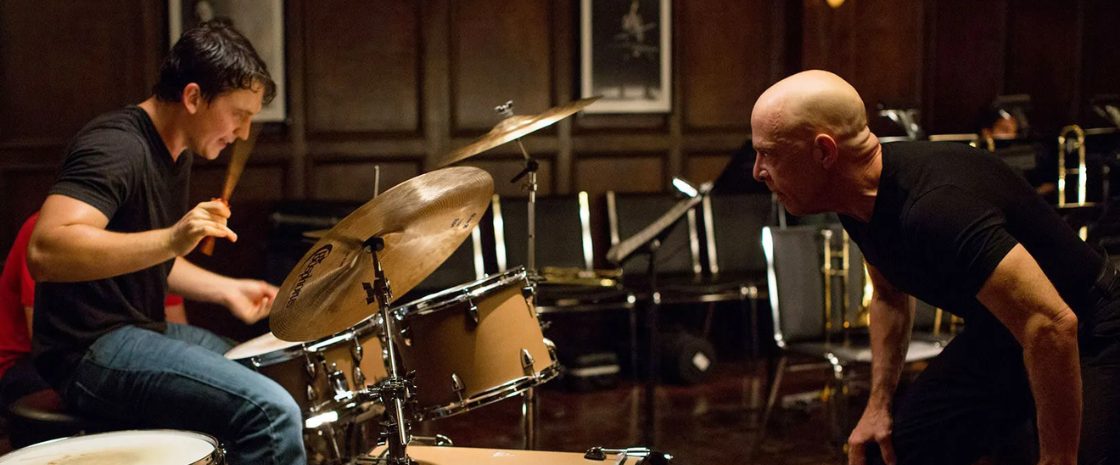 A scene from the movie 'Whiplash' featuring Miles Teller as Andrew Neiman intensely playing the drums, while J.K. Simmons as Terence Fletcher leans in, watching closely in a rehearsal room setting.