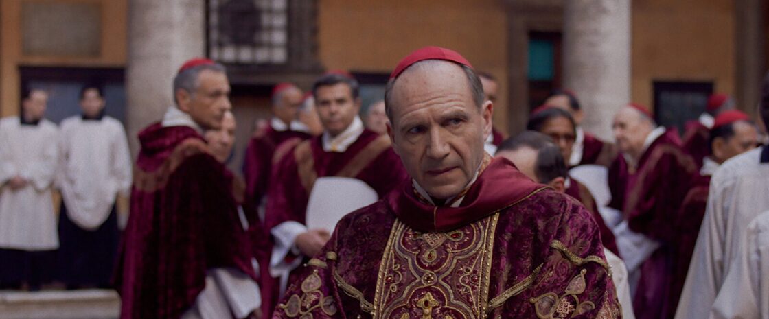 Ralph Fiennes as Cardinal Thomas Lawrence in Conclave (2024), surrounded by fellow cardinals in a Vatican courtyard.