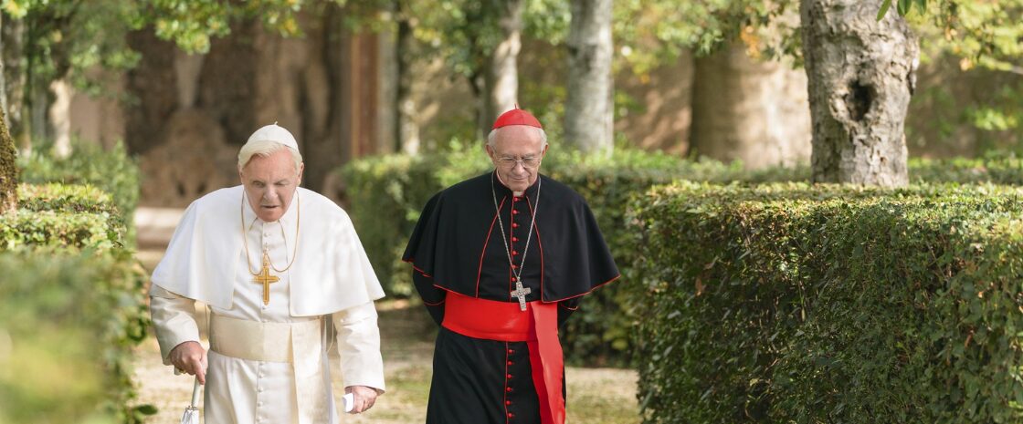 Anthony Hopkins as Pope Benedict XVI and Jonathan Pryce as Cardinal Jorge Bergoglio walking through a garden in a scene from The Two Popes.