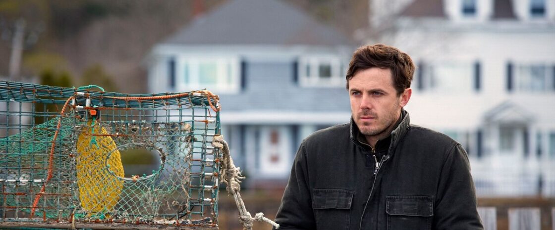 Casey Affleck as Lee Chandler standing near a lobster trap with coastal homes in the background in Manchester by the Sea.