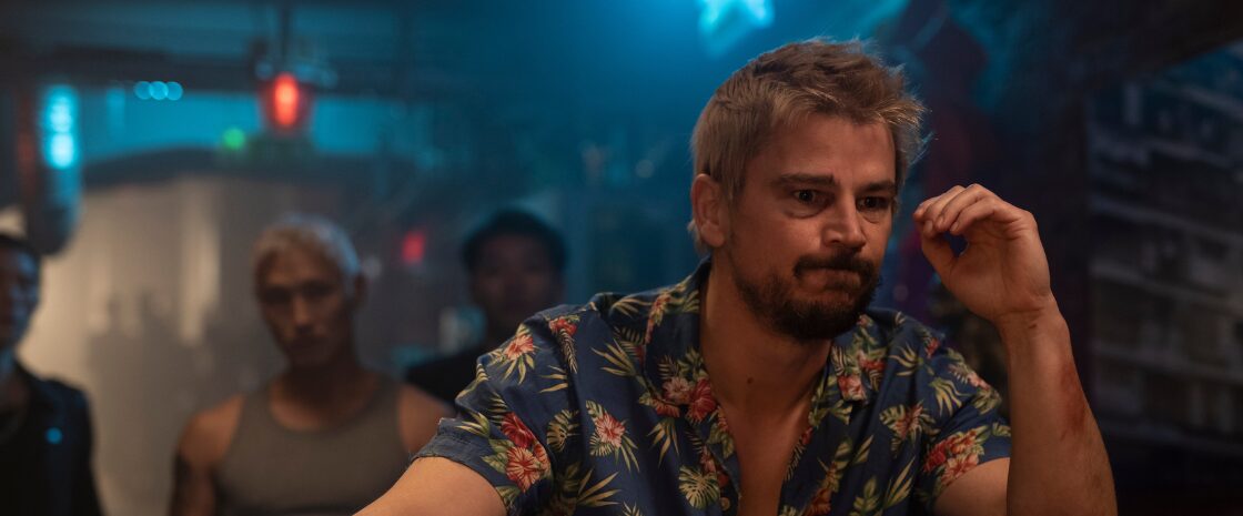 Josh Hartnett as Lucas Reyes in Fight or Flight, sitting tensely at a bar in a dimly lit, neon-blue scene.